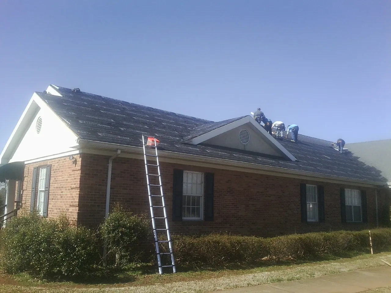 Crew tearing shingles off a brick ranch roof with a ladder set — Emergency Roof Repair work by our crew in Gansevoort