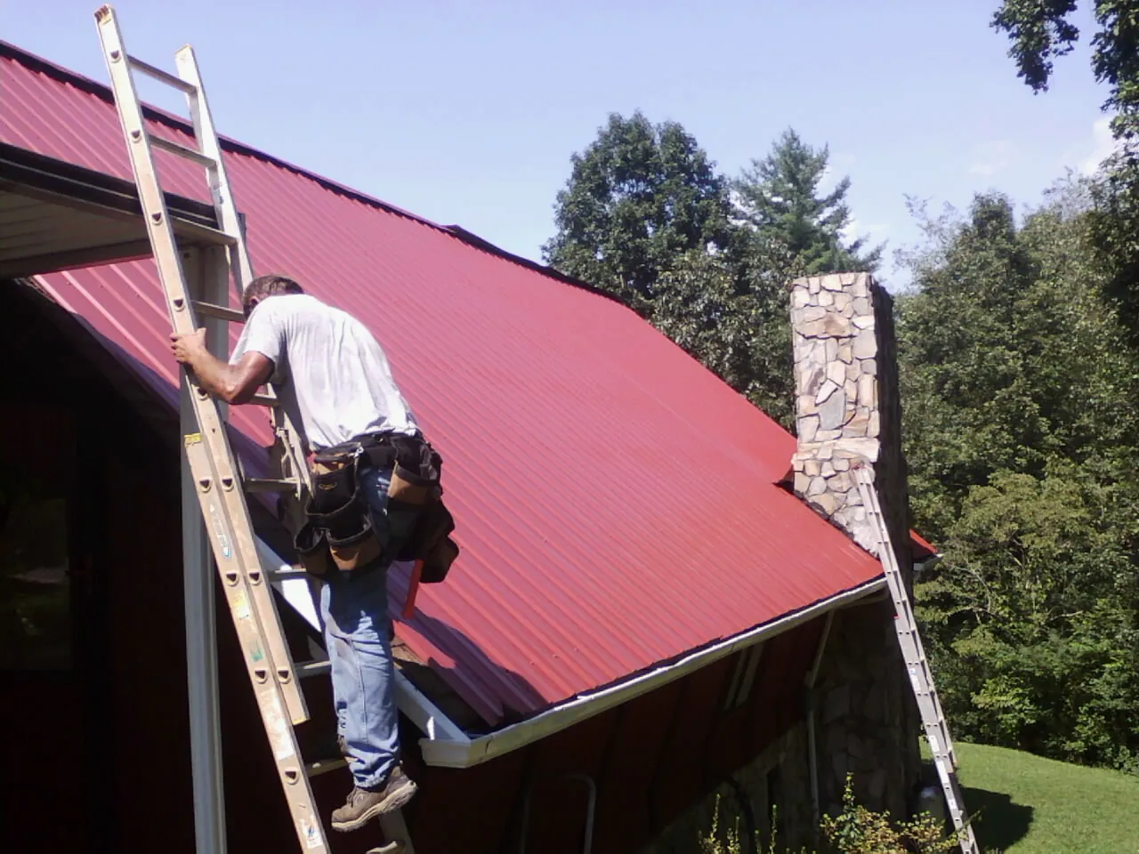 Roofer on a ladder beside a red metal roof and stone chimney — Soffit & Fascia Repair by Roofing Brothers in Gansevoort